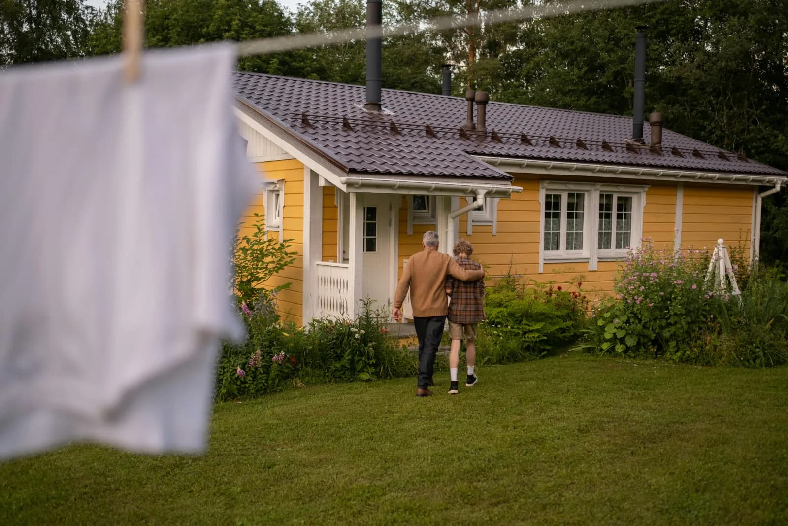 To personer går arm i arm mot et gult trehus i hagen i gyllent kveldslys