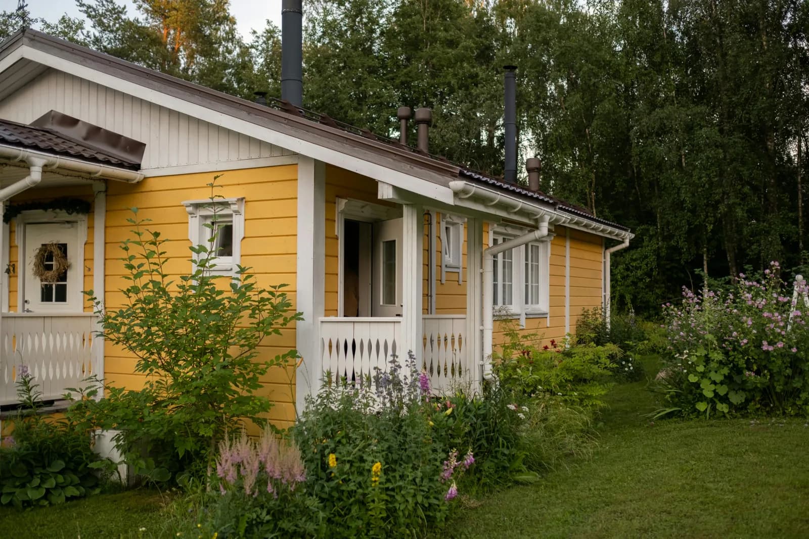 Gult trehus med hvit veranda og frodig blomsterhage foran skogen