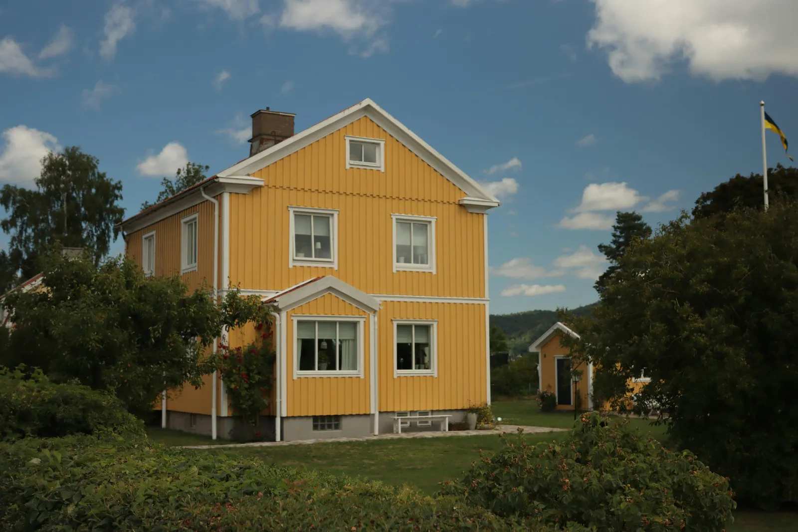 Gult toetasjes trehus med hvite vinduskarmer i en frodig hage under blå himmel